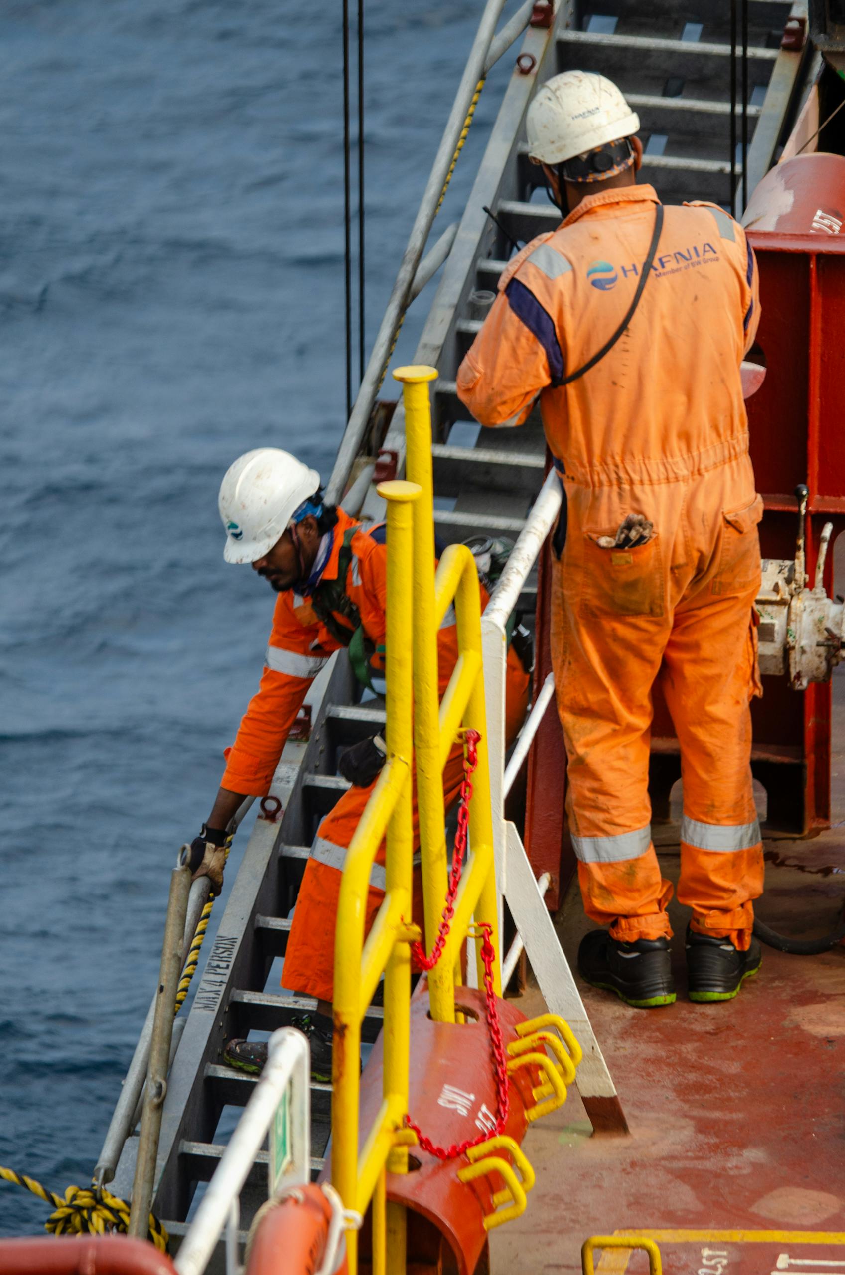 Two offshore workers in orange gear on a vessel near open sea performing maintenance.